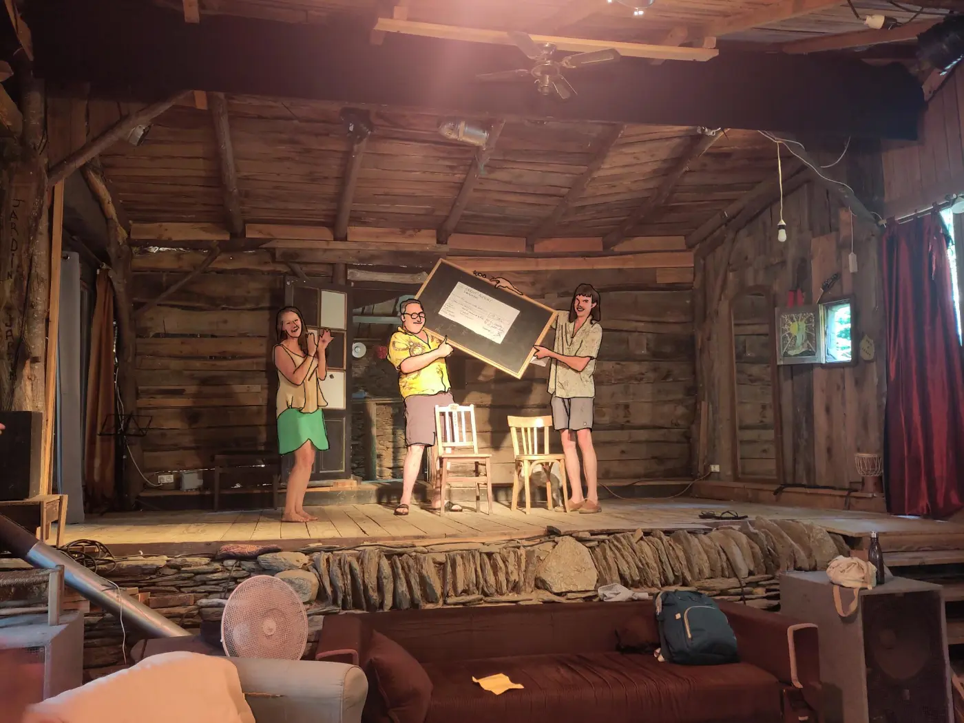 Sur la scène en bois du théâtre, trois personnes souriantes présentent une grande ardoise où une feuille est affichée.