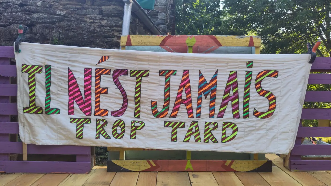 La banderole cousue « Il n'est jamais trop tard », lettres rayées multicolores sur drap blanc, accrochée à une palissade violette.